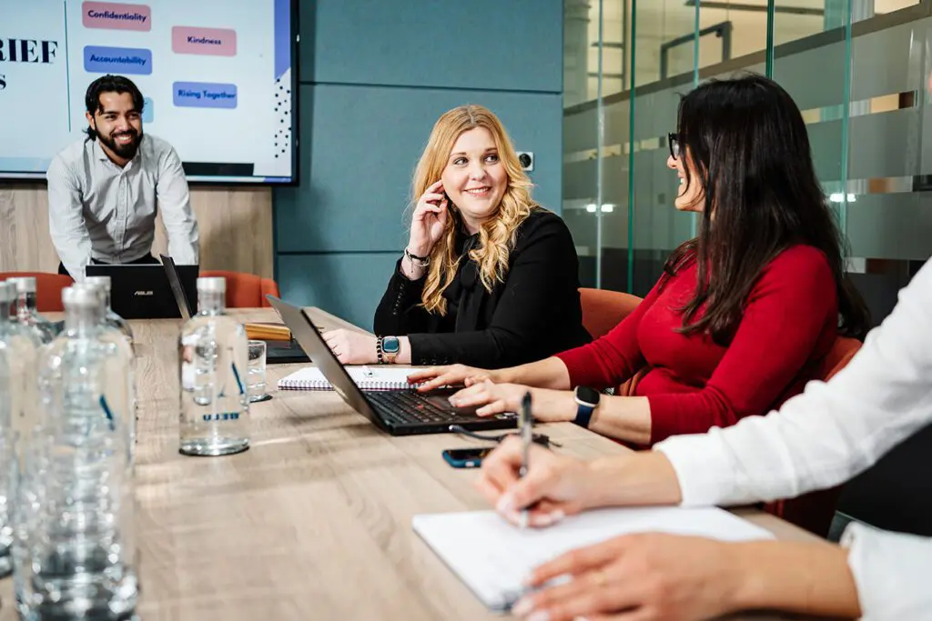 Two female workers sat around on a long table, dressed in red and black, discussing work whilst a male presents a presentation in the background.