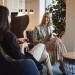 Two professionally dressed women converse in a stylish, modern lounge.